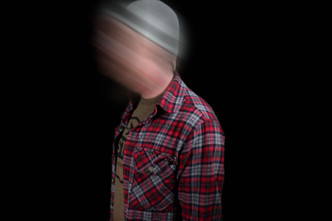 Man wearing a flannel shirt, green shirt, and beanie in a dark room.