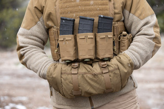 A man, wearing a merino wool jacket and body armor, standing outside with his hands tucked inside of a hand-warmer.